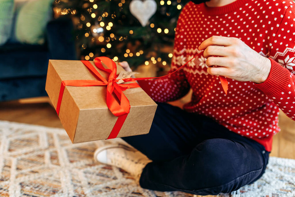 young man is holding a Christmas present with a red ribbon. Close-up, Gift opening - Фото, изображение