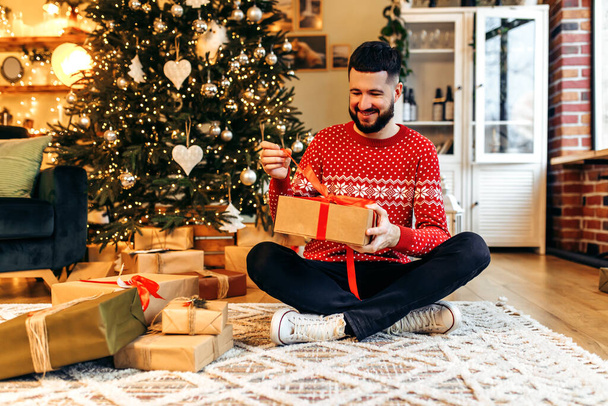 Smiling young man sitting near the Christmas tree, with a Christmas present at home, New Years celebration, Merry Christmas - Foto, afbeelding