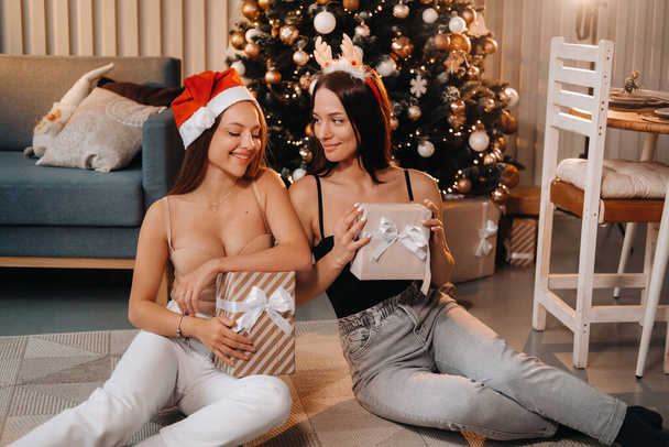 Дві красиві молоді дівчата з різдвяними подарунками в руках сидять біля ялинки в будинку.
. - Фото, зображення