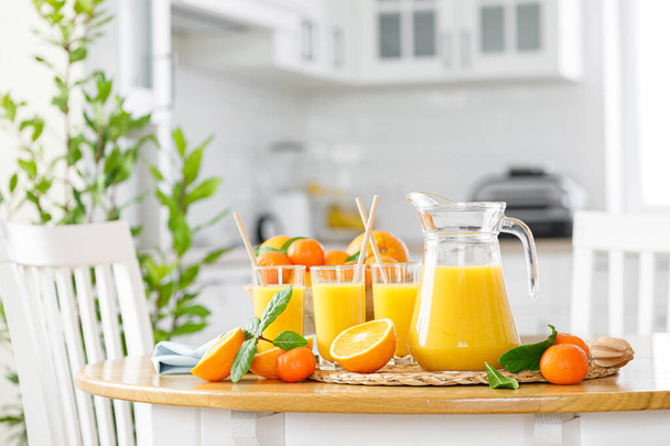 Freshly squeezed orange juice in a glass jug, glasses and fresh fruits with leaves on the table against the background of modern white kitchen, healthy drink for breakfast, vitamin c concept, front view - Фото, изображение