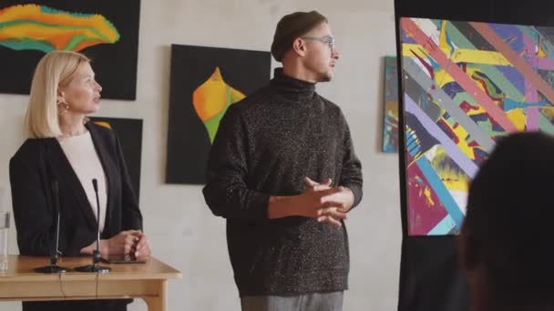 Young trendy male artist speaking with female auctioneer and giving presentation about his abstract painting to audience during art auction in gallery - Footage, Video