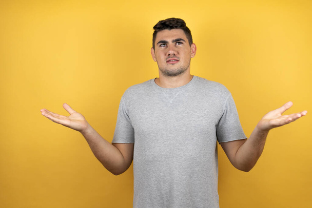 Young handsome man wearing a casual t-shirt over isolated yellow background clueless and confused expression with arms and hands raised - Photo, Image