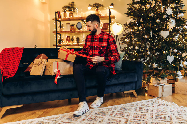 счастливый молодой человек, открывающий рождественские подарки дома на фоне украшенной елки, счастливого Рождества и Нового года - Фото, изображение