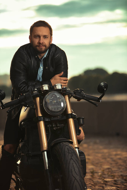 Modischer Biker in schwarzer Lederjacke sitzt auf seinem Motorrad und blickt in die Kamera. - Foto, Bild