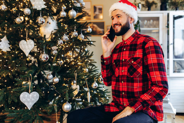 Smiling young man in Santa Claus hat talking on cell phone at home against background of Christmas tree - Φωτογραφία, εικόνα