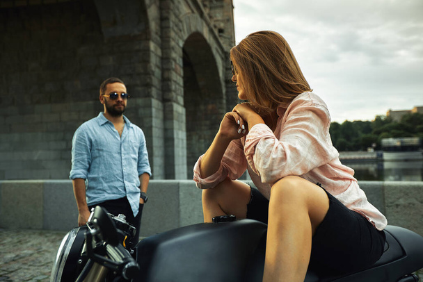 Cita romántica en moto. Mujer joven se sienta en una motocicleta y mira al hombre que viene a ella. Pareja enamorada de la puesta de sol bajo el puente en la ciudad - Foto, Imagen