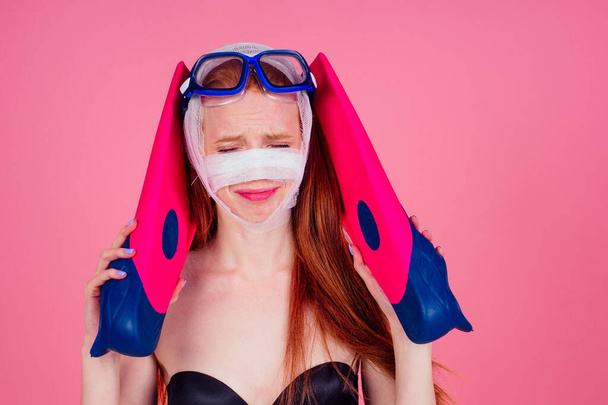redhaired ginger woman with a bandaged face holding flippers and looking sad in studio pink background - Φωτογραφία, εικόνα
