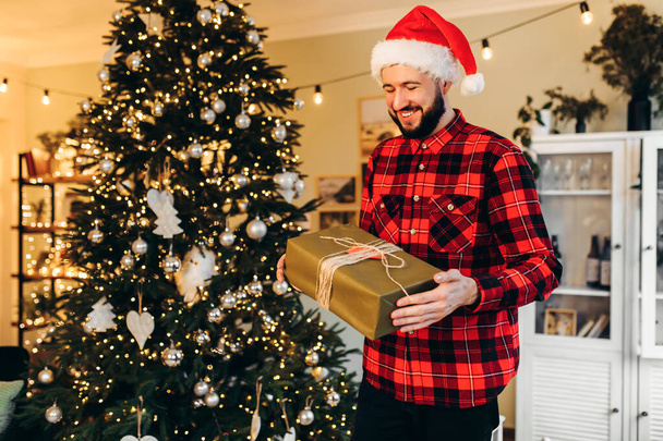 Smiling stylish young man in Santa Claus hat with Christmas present enjoying Merry Christmas at home, near decorated Christmas tree, Merry Christmas and Happy New Year - Foto, Bild