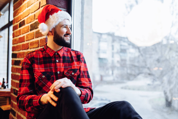 Attractive christmas man wearing santa claus hat sits on windowsill and looks out the window enjoying christmas in stylish home - Photo, image