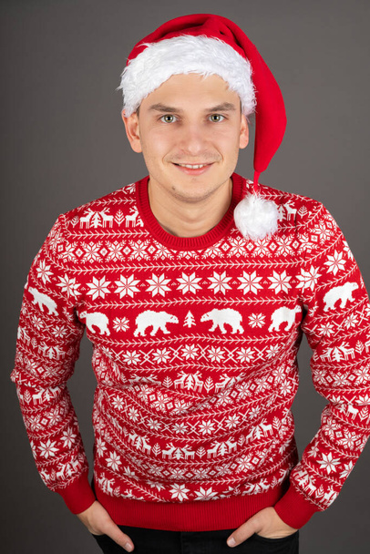 Handsome young man wearing Santa hat and red Christmas style pullover on grey background - Φωτογραφία, εικόνα