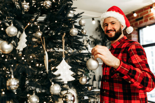 Young happy man in Santa Claus hat, decorates the Christmas tree with toys, prepares for the Christmas holiday at home, Merry Christmas - Fotografie, Obrázek