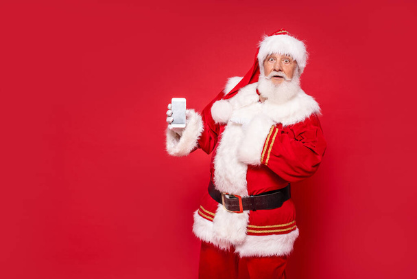 Shocked Santa Claus showing mobile phone screen, pointing on smartphone. Red xmas studio background. Christmas sale. - Foto, Imagem