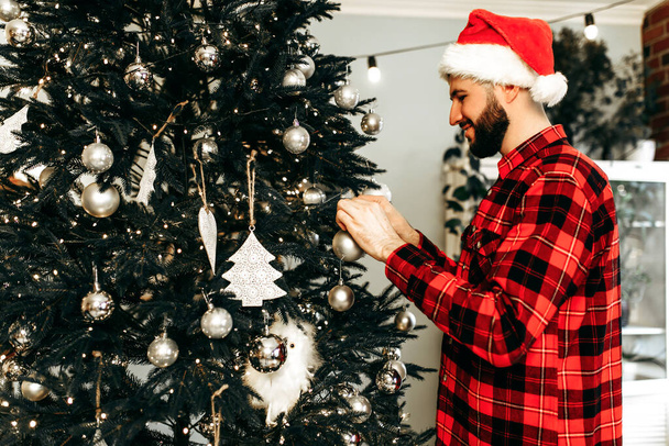 Young happy man in Santa Claus hat, decorates the Christmas tree with toys, prepares for the Christmas holiday at home, Merry Christmas - Photo, image