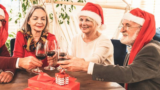 Happy senior friends celebrating Christmas together, wearing red Santa Claus cap, toast with red wine at the home party. Elderly people lifestyle and holidays concept. - Photo, image