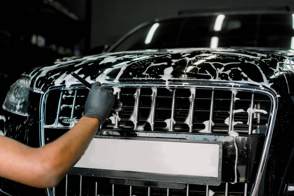 Car detailing wash. Manual cleaning with soap at car wash. Cropped image of hand of male worker in rubber glove washing car radiator grille of luxury black car with special brush and cleansing foam - Foto, afbeelding