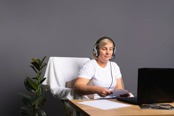Pandemic and remote work. A woman at home in an armchair works with office papers in front of a computer in headphones. High quality photo - Foto, Imagem
