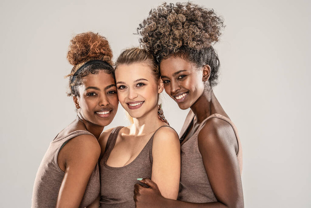Portrait of three beautiful, smiling multicultural young women standing close to each other, looking at camera. Beauty and spa concept. Multi ethnic group of happy female friends.Studio shot. - Фото, зображення
