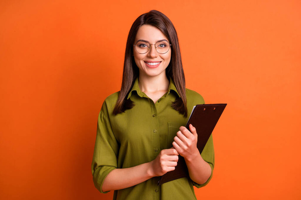 Foto retrato de trabalhadora segurando prancheta sorrindo vestindo óculos camisa verde isolado no fundo cor de laranja vibrante - Foto, Imagem
