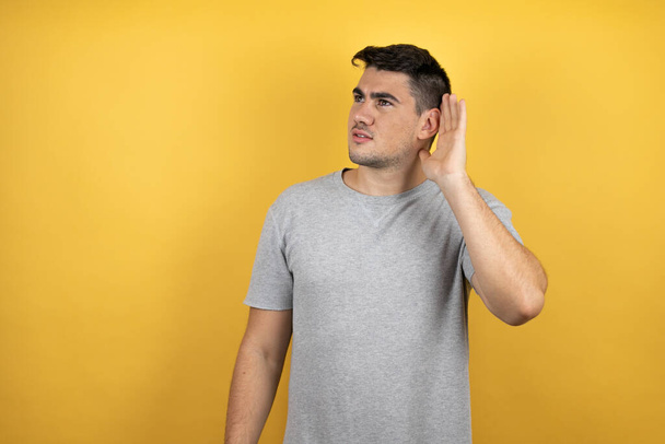 Joven hombre guapo con una camiseta casual sobre fondo amarillo aislado sorprendido con la mano sobre el oído escuchando un rumor o chismes - Foto, imagen