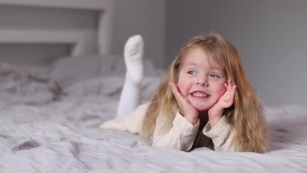 heureuse jolie petite fille blonde en robe beige couchée sur un lit dans une chambre grise. routine du matin, enfance, tout-petit, fille. Vidéos en Full HD - Séquence, vidéo