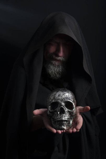 A man in the form of a sorcerer holds a human skull in his palms, magical deeds - Foto, imagen