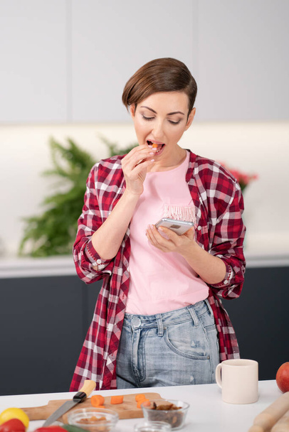 Housewife checking email or texting or posting photos of food for her social media using her smartphone while cooking fresh salad wearing a plaid shirt. Healthy food leaving - vegan concept - Fotoğraf, Görsel