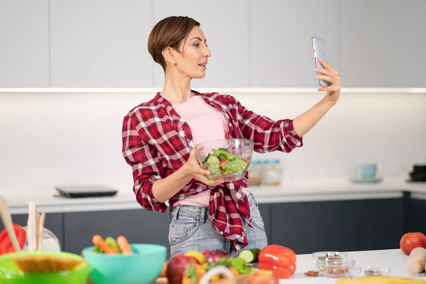 Pretty housewife taking selfie or making a video call using her smartphone while cooking fresh salad wearing a plaid shirt with a bob hair style. Healthy food leaving - vegan concept - Zdjęcie, obraz