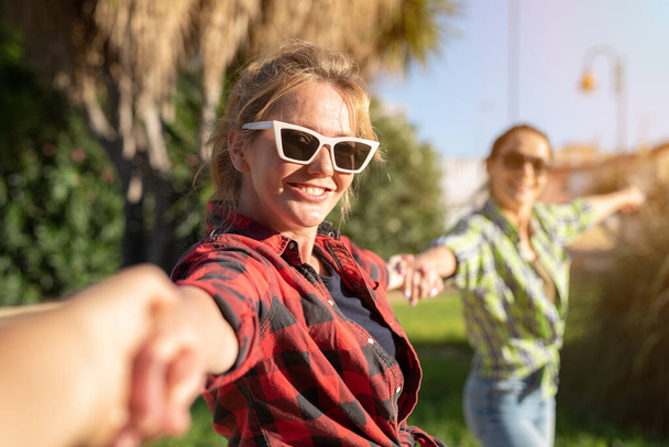 Mooie jonge blanke vrouw met een zonnebril die uitnodigend met een vrolijke glimlach langs de hand trekt als haar vriendin op de achtergrond. Zonnige zomerdag. - Foto, afbeelding