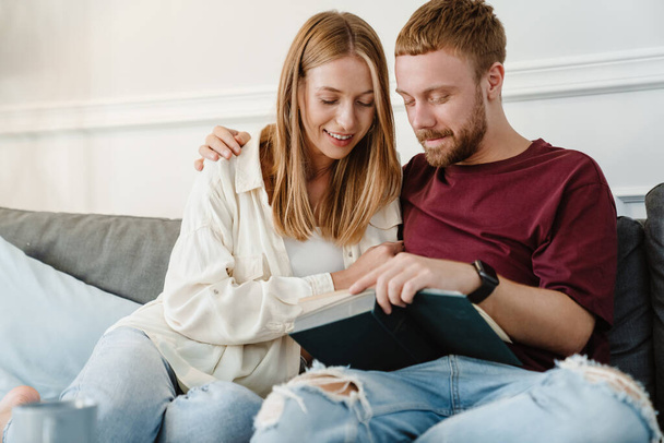 Photo of happy romantic couple hugging and reading book while sitting on couch at living room - Foto, Bild
