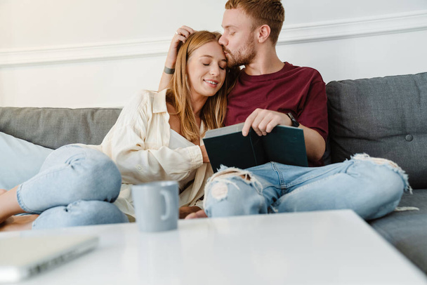 Photo of happy romantic couple kissing and reading book while sitting on couch at living room - Foto, imagen