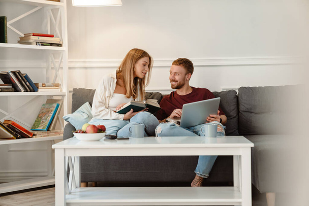 Зображення імбиру задоволена пара читає книгу і використовує ноутбук, сидячи на дивані вдома
 - Фото, зображення