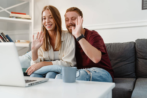 Image of ginger joyful couple waving hand and using laptop while sitting on sofa at home - Foto, Imagen