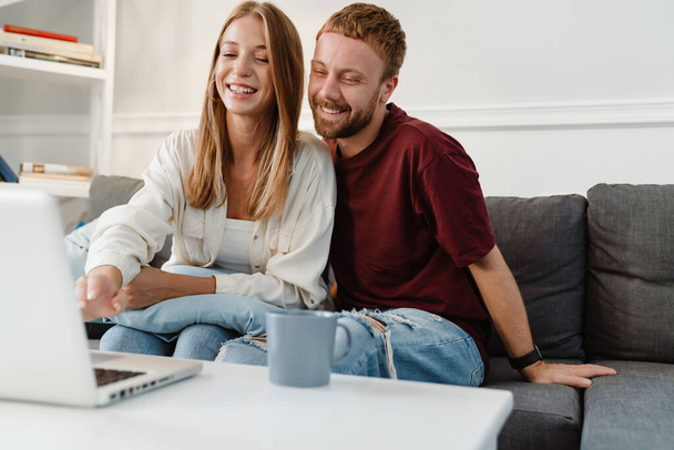 Image of ginger joyful couple laughing and using laptop while sitting on sofa at home - Foto, Imagen