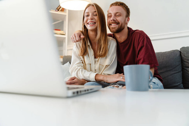 Image of ginger joyful couple laughing and using laptop while sitting on sofa at home - Фото, зображення