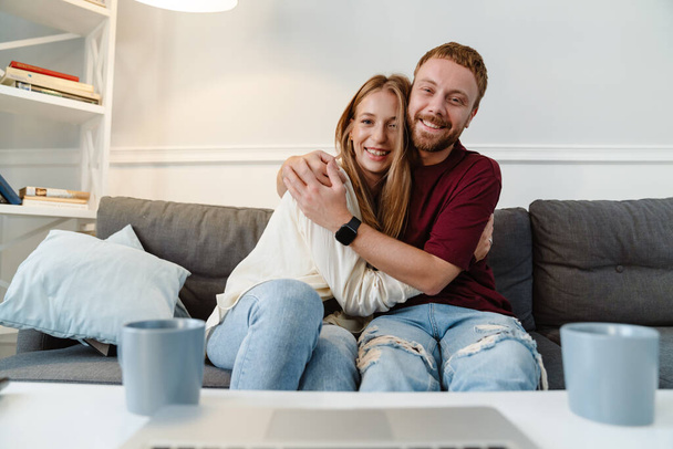 Image of ginger joyful couple hugging and using laptop while sitting on sofa at home - Fotoğraf, Görsel