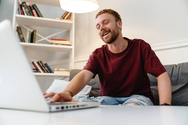 Image of ginger joyful guy laughing and using laptop while sitting on sofa at home - Zdjęcie, obraz