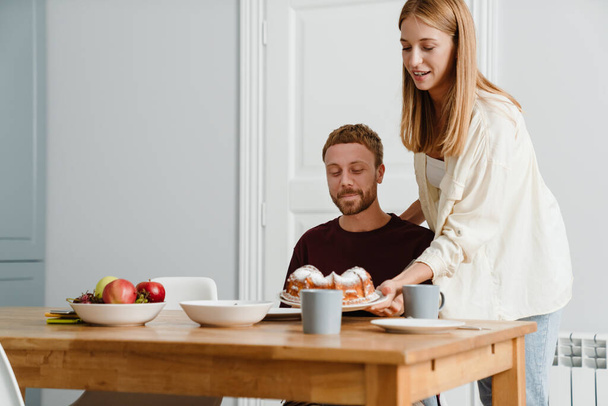 Image of ginger joyful couple drinking coffee with pie while sitting at table in home - Fotografie, Obrázek