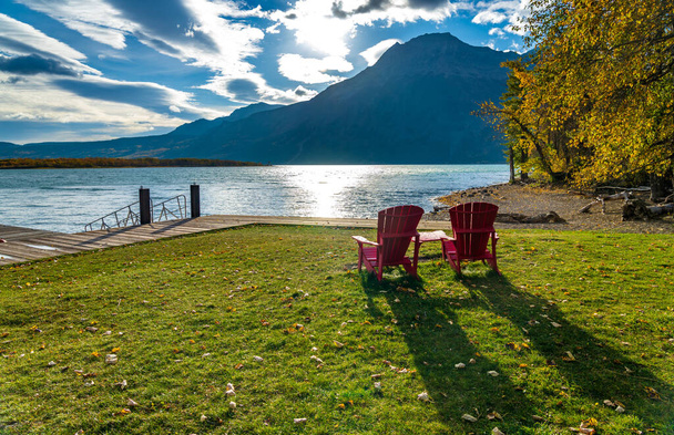 Red chair look over Waterton Lake Marina Point in autumn foliage season sunny morning. Blue sky with colourful clouds reflect on the lake water surface. Waterton Lakes National Park, Alberta, Canada. - Photo, Image