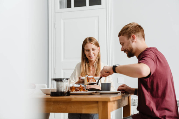Image of ginger joyful couple drinking coffee with pie while sitting at table in home - Foto, Imagem