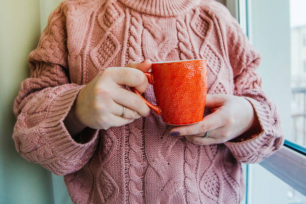Ένα κορίτσι με πουλόβερ κρατάει ένα πορτοκαλί φλιτζάνι καφέ ένα ηλιόλουστο πρωινό. - Φωτογραφία, εικόνα