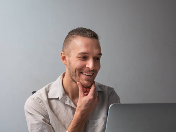 Hombre caucásico está agarrando su barbilla, sonriendo frente a la computadora, hablando en videollamada en cuarentena por la pandemia del coronavirus - Foto, imagen