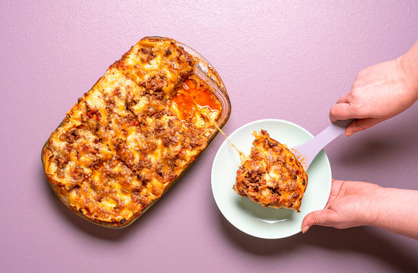Above view with woman hands serving on a plate a portion of freshly-baked lasagna. Delicious homemade lasagna top view on a purple background. - Photo, Image