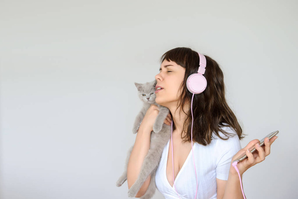 cerca de la foto de una morena feliz con auriculares rosados escuchando la música mientras sostiene un gatito gris en sus brazos - Foto, imagen