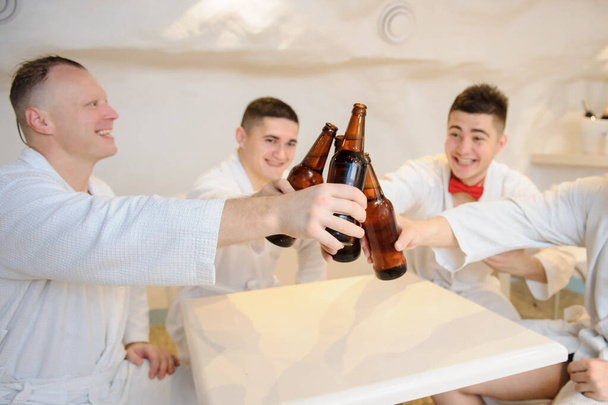 白いガウンを着た4人の男がテーブルの周りに座ってサウナの後でビールを飲んでる写真 - 写真・画像