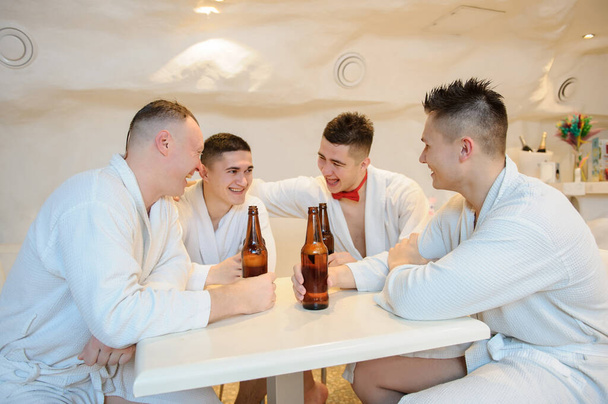 close up photo of 4 men in white gowns sitting around a table and drinking beer after sauna procedures - Photo, image