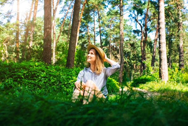 Hipster lány szalmakalapban ül az erdőben. Vándorlási vágy koncepció. Utazási ötletek. Gyönyörű nő a természetben a fűben. Nyári rezgések. - Fotó, kép