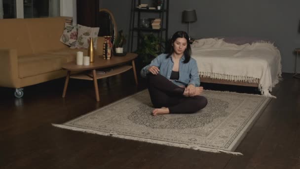 Relaxado jovem mulher asiática em sportswear está meditando na posição de lótus sentado no chão com os olhos fechados em casa desfrutando de meditação com as mãos em namaste. - Filmagem, Vídeo