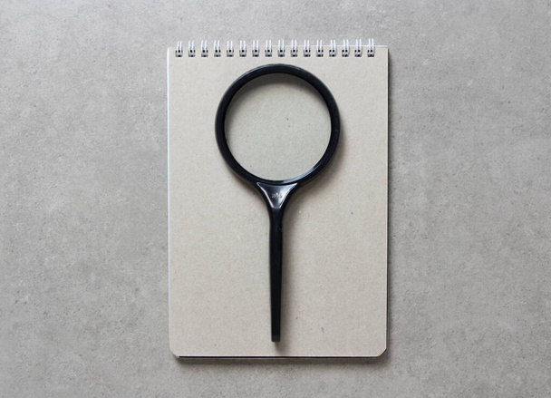 A spring notebook with a sheet of craft paper A5 with black magnifying glass on light grey concrete background. Concept of new idea, business plan and strategy, development and implementation of content. Empty space for text. - Photo, Image