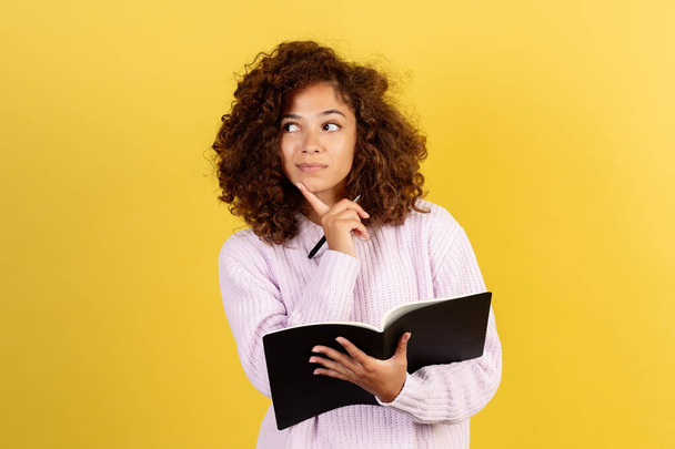 Ponder young afro american student girl looking aside, making thoughtful face, thinking about exam test and education in college, standing on yellow copy space background - 写真・画像