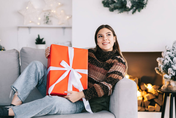 Happy caucasian woman holding big Christmas present while sitting on a sofa at home near Christmas tree. High quality photo - Foto, Imagen